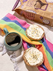 Bread SRSLY gluten-free sourdough sandwich rolls topped with cream cheese and everything bagel seasoning next to a cup of hot coffee.